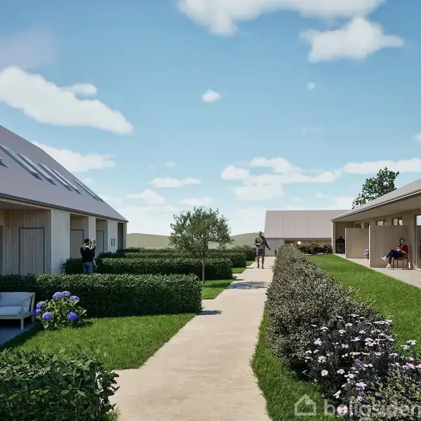 To rækker moderne huse står på hver side af en græsplæne med blomsterbede og en sti i midten, omgivet af en klar blå himmel med spredte skyer.