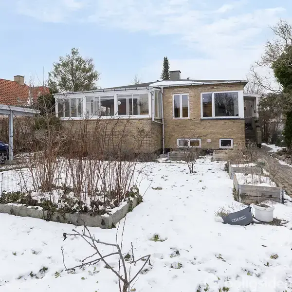 Et enkeltfamiliehus står i en snedækket have med terrasserede bede. En carport med en blå bil er til venstre, og en stensti fører til huset.