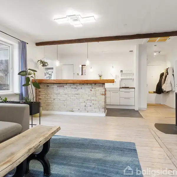 En stue med en grå sofa og et træbord står foran et åbent køkken med en bar lavet af hvide mursten. Brændeovn i hjørnet til højre.