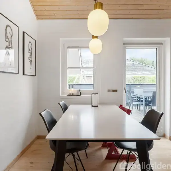 Et hvidt spisebord med sorte stole står i et lyst rum med træloft. To lamper hænger over bordet, og der er billeder på væggen samt udsigt til en veranda.