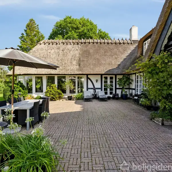Et stråtækt hus står i en solrig gårdhave med flisebelægning. En parasol dækker et udendørs spisebord foran huset, omgivet af frodig vegetation og klar blå himmel.