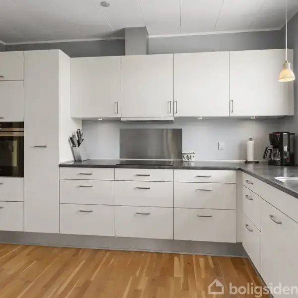 Et moderne køkken med hvide skabe og skuffer, indbygget ovn og kogeplade. En knivblok står ved siden af vasken. Vindue med udsigt og trælaminatgulv.