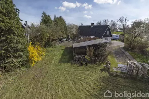 En gammel lade står stille på en grøn græsplæne omgivet af træer og buske i en landlig have. Ved siden af laderen er der et traktorskurt.