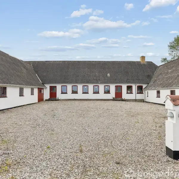 Et firlænget bindingsværkshus med stråtag omkranser en grusbelagt gårdsplads. Huset har hvide vægge med røde døre og vinduer. Til venstre ses en postkasse på en hvid muret søjle.
