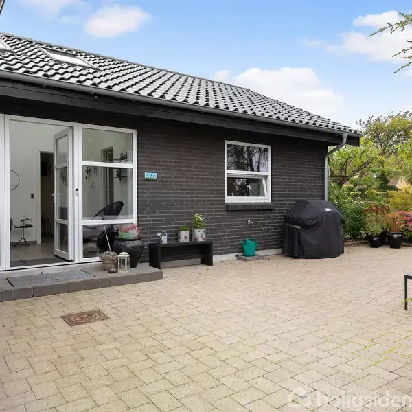 Et sortmuret hus med åbne glasdøre fører ud til en flisebelagt terrasse. Omkring terrassen er der blomsterrige planter og havemøbler.