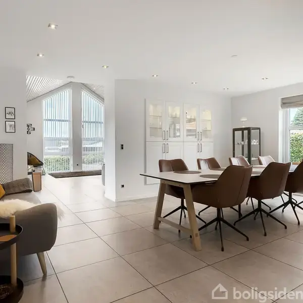 Spisebord med otte stole står i en moderne spisestue. Store flisebelagte gulve og vinduer giver lys. Sofa og billeder på væggen til venstre. Ingen tekst tilstede.
