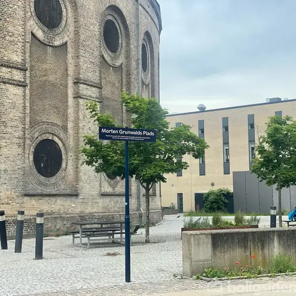 Rund bygning med runde vinduer står ved siden af et skilt med teksten "Morten Grunwalds Plads". Personer sidder ved et bord på en brolagt plads med træer og bygninger omkring.