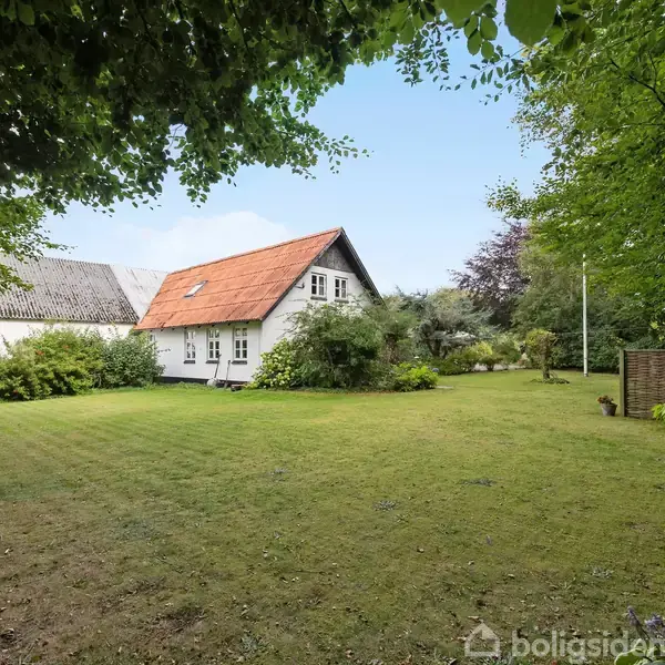 Huset med rødt tag står stille i en grøn have omkranset af tætte træer og buske, med en velplejet græsplæne foran.