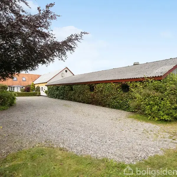 En lang bygning dækket af vedbend står stille ved en grusvej. I baggrunden ses flere huse med røde tage, omgivet af træer og buske.