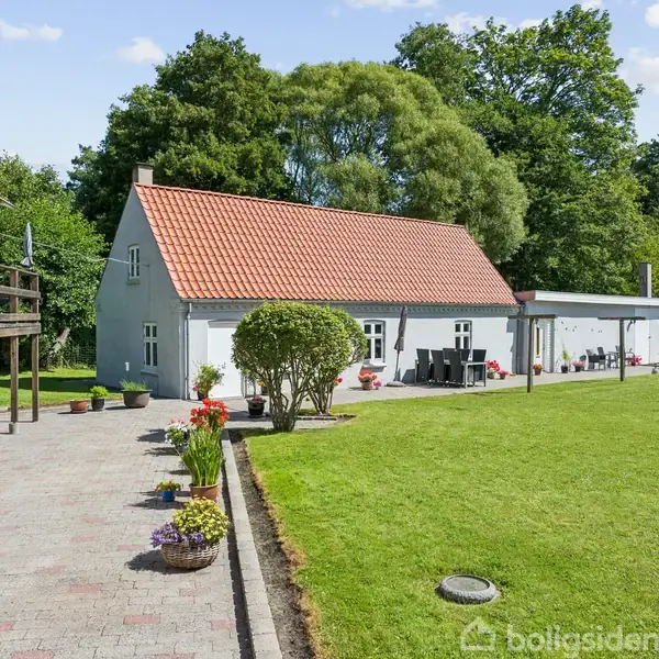 Et hvidt hus med rødt tag står ved siden af en velplejet græsplæne og en brostensbelagt sti med blomsterkrukker. Bagved ses træer og en klar blå himmel.