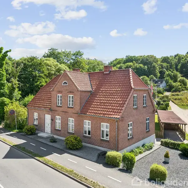 Et rødt murstenhus med rødt tegltag stående ved en vej omgivet af grønne træer og buske. Ved siden af huset er en overdækket carport. Skov fylder baggrunden.