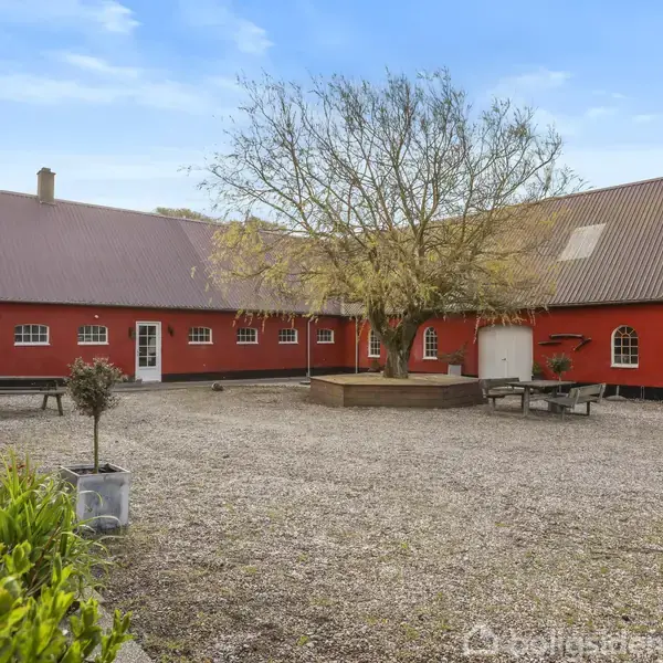 En rød bygning med buede vinduer omkranser en gårdsplads. Træ i midten, omgivet af grus. Picnicborde står ved siden af træet i den åbne plads.