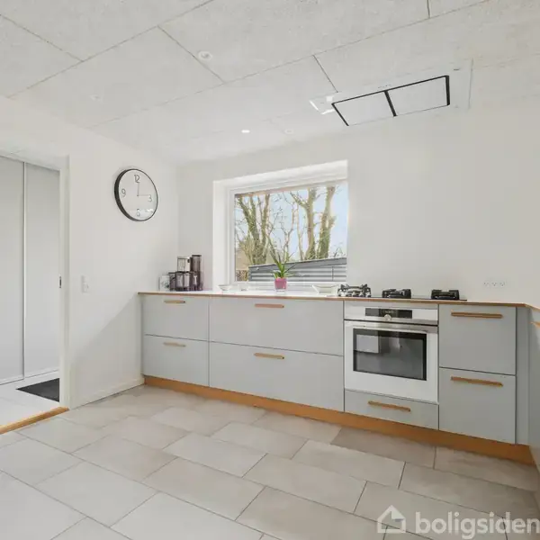 Et lyst køkken med grå skabe og hvide vægge har en indbygget ovn og gaskogeplader. En væg har et vindue med udsigt til træer. Der er et analogt vægur på væggen.