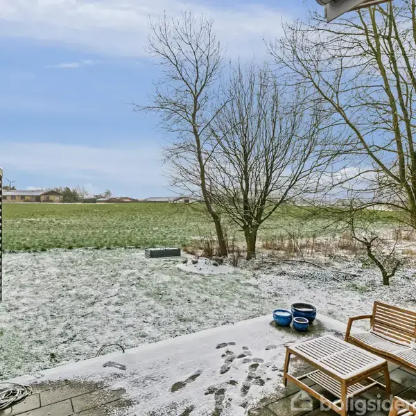 En terrasse med sneklædt flisebelægning. Bænke udenfor huset med udsigt til et åbent, snedækket landskab. Træer står nøgne under en blå himmel.