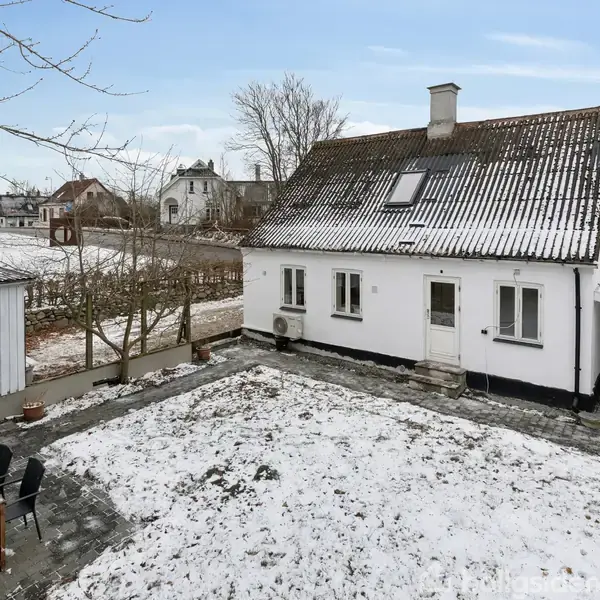 Et gammelt hvidt hus med skråt tag står i en snedækket gårdhave. En terrasse med sorte stole og et bord står til venstre. Naboer ses i baggrunden.