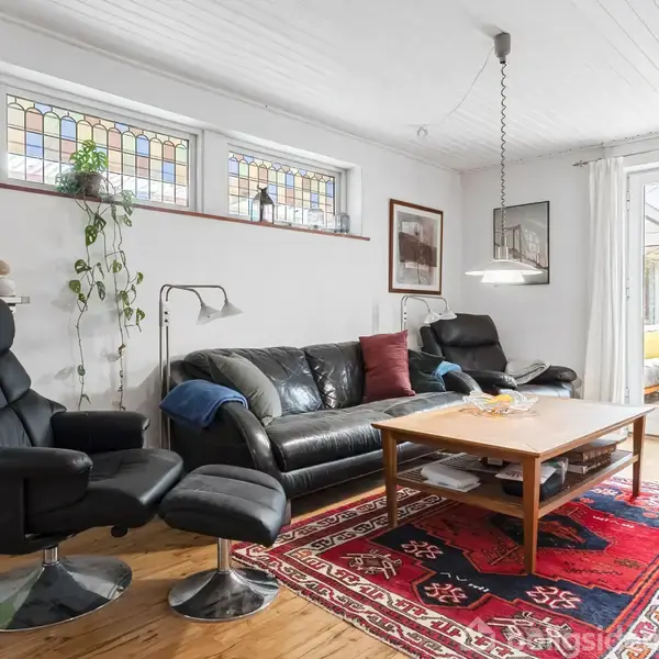 En stue indeholder en sort lædersofa med puder, en sort lænestol med skammel, et træbord på et farverigt tæppe, og en hylde med planter og bamser. Der er store vinduer med farvede glasindlæg.