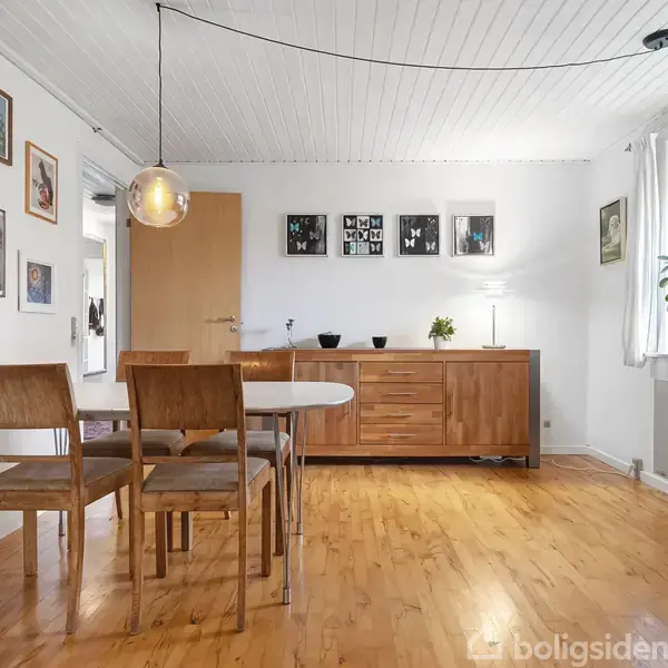 Et spisebord med fire træstole står i et lyst rum. En kommode og vægkunst pryder væggen. Et vindue med planter tilføjer naturligt lys.