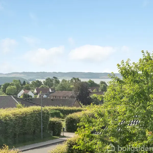 Udsigt over boligområde med huse omgivet af hække og træer. I baggrunden ses åbne marker under en klar blå himmel. Grønt løv dominerer forgrunden.