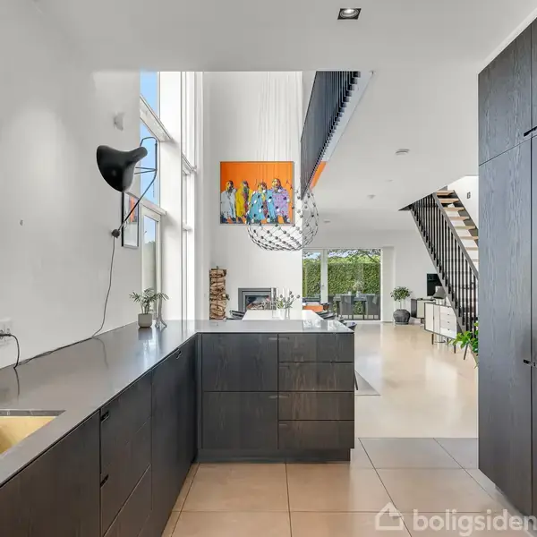 Et moderne køkken har mørke skabe og vask, set i aktion i en rummelig, lys stue med højt til loftet, store vinduer, en kunstplakat og en trappe i baggrunden.