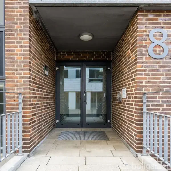 Indgangsdør til en bygning med husnummeret 8A, lavet af glas og metal. Omgivet af murstensvægge, placeret i en moderne arkitektonisk ramme med metalrækværk på begge sider.