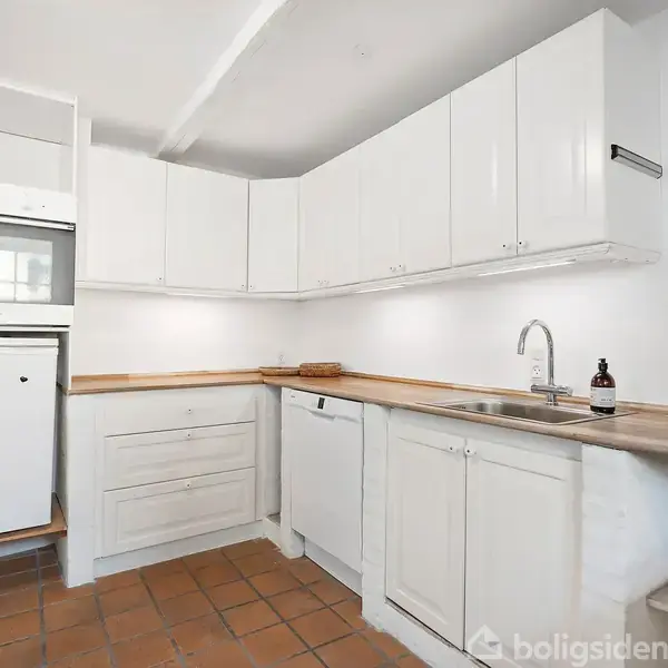 Et køkken med hvide skabe hænger over en træbordplade. En vask med en vandhane ses, fliser dækker gulvet, og der er en opvaskemaskine samt en ovn og et køleskab.