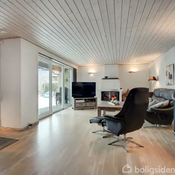 Stue med lænestol og sofa foran fladskærms-TV og brændeovn. Store vinduer til venstre giver udsigt til have, mens vægge og loft er hvidmalet.