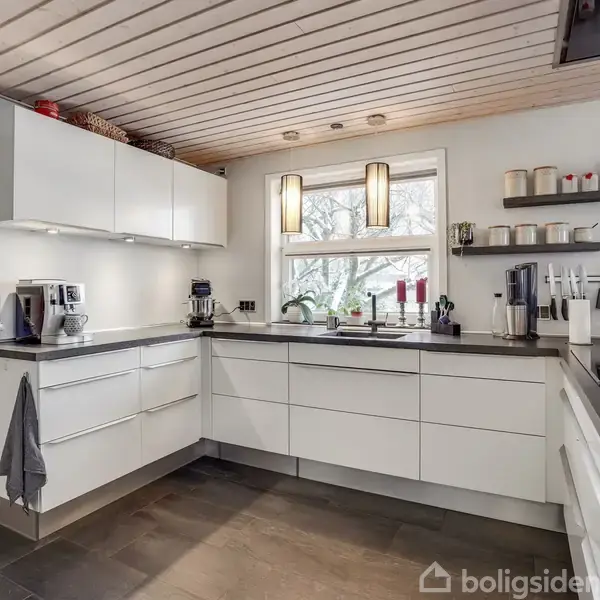 Et langt spisebord med stole står i en moderne spisestue med lyse trævægge. Et stort, rektangulært lysarmatur hænger over bordet, ved siden af åbent køkken og vindue.