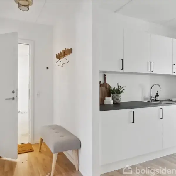 Dør åben til modernistisk køkken med hvide skabe og grå bordplade; planter og skærebræt pynter. Træbænk langs væg med knagerække ovenover. Lyst træparketgulv.