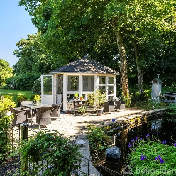 En pavillon står på en stenbelagt terrasse omgivet af havemøbler. Ved siden af findes en dam med blomster. Omgivelserne består af græsplæner og træer i et frodigt haveområde.