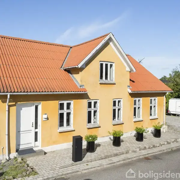 Et gult hus med rødt tegltag, står foran en vej. Fire sorte krukker med planter står foran huset. En hvid campingvogn er parkeret ved siden af på grus.
