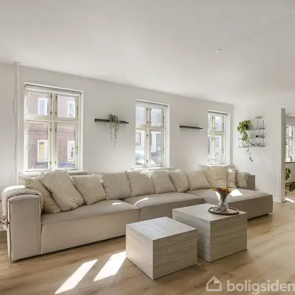 Sofa med lyse puder står mod en væg med tre vinduer. I forgrunden ses et sofabord med blomster. Rummet er lyst og indrettet med minimalistiske detaljer og planter.