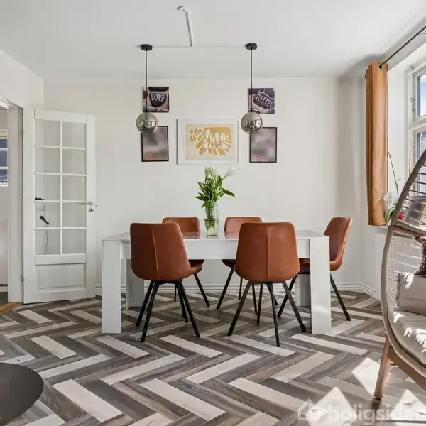 Spisebord med fire brune stole og en vase med blomster. Over det hænger to lamper. Væggene har kunstværker, og gulvet er zebrastribet. Ved siden af er en hængestol ved vinduet.