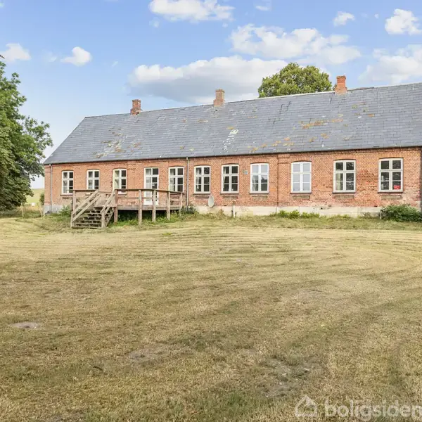 En lang, ældre rød murstensbygning med mange vinduer står i en åben, velplejet græsplæne omgivet af træer. Trætrappe fører op til indgangsdør. Skyer ses på himlen.