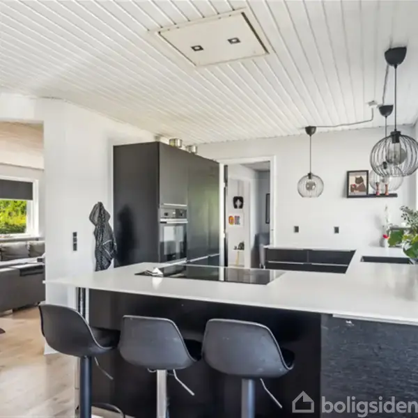 Sort køkkenbord med tre barstole, integreret kogeplade og overhængende lamper. I baggrunden ses en stue med sofaer. Blomsterpotte på bordet. Tekst på væg: "Livet er ikke..."