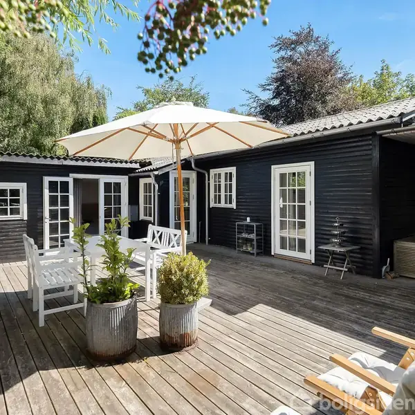 Sort træhus står på en solrig terrasse, hvor et hvidt bord og stole er dækket af en stor parasol. Omkring det er frodig grøn vegetation.