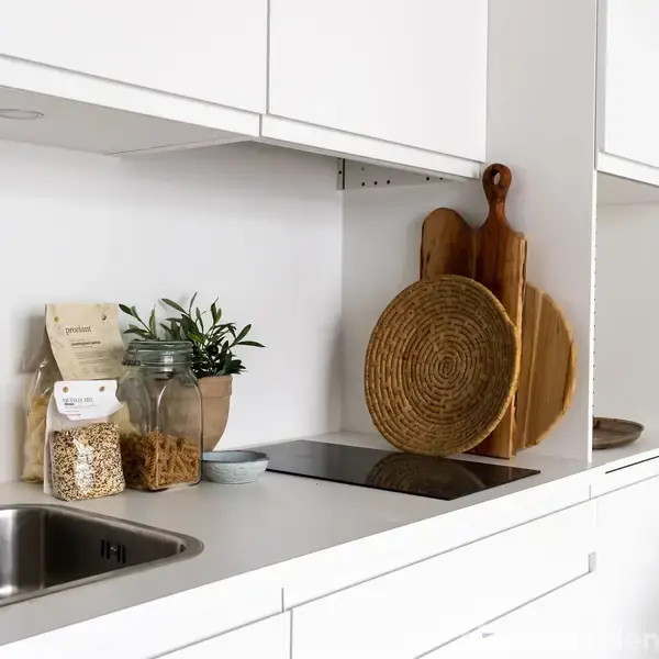 Hvidt køkken indrettet med vask, komfur og dekorative skærebrætter. På bordpladen står krukker med ingredienser og en lille plante. Lyse og minimalistiske omgivelser.