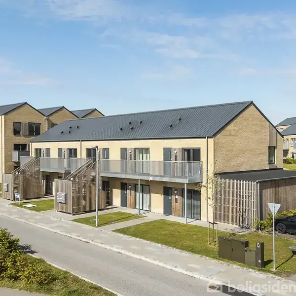 To-etagers rækkehus med murstensfacade og altaner; omgivet af vej, fortov og grønne områder; biler parkeret i nærheden under en blå himmel.