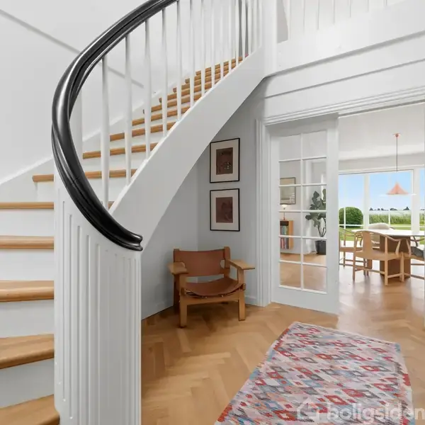 Et moderne køkken med en stor, central køkkenø prydet med frugtskål. To hængelamper hænger over øen. Omkring er der hvide skabe og træparketgulv i et lyst rum.