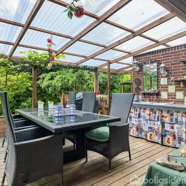 Udendørs terrasse med spisebord og seks stole under et gennemsigtigt tag. Omringet af frodige planter og mure med dekoreret bordplade og opsat små pyntegenstande. Afslappet atmosfære.