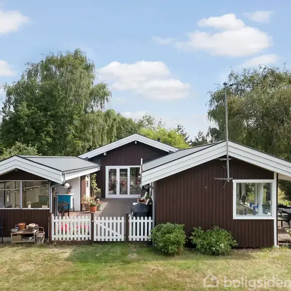 Brun træhytte står stille omgivet af grønne buske og træer i en velplejet have med blå himmel og hvide skyer. Der er en lille terrasse og hvidt hegn.