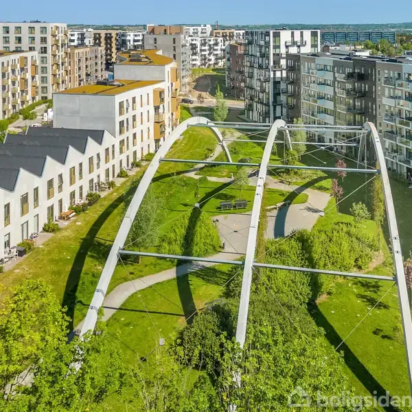 Et grønt område med stier er omgivet af moderne boligblokke og rækkehuse. Store, buede metalstrukturer krydser over parken. Sollys belyser området fra en klar blå himmel.