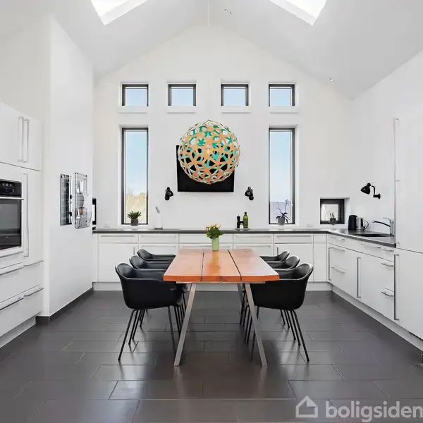 Et lyst køkken med et langt træbord omgivet af sorte stole. Kunst og vinduer bagved tilføjer dekorativt præg. Køkkenudstyr og skuffer omkranser det hele, hvilket skaber et moderne miljø.
