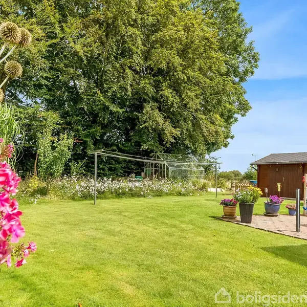 Græsplæne med blomster i forgrunden. Et tørrestativ står i midten. I baggrunden ses et træ og et lille træhus med blomsterkrukker. Klar himmel over det hele.