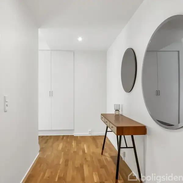 Spejl hænger på en hvid væg, reflekterer lys fra gangen. Under det står et smalt træbord med sorte ben. Trægulv og indbyggede skabe fuldender det minimalistiske rumindtryk.