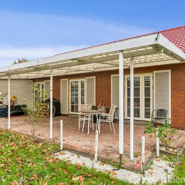 En åben terrasse med hvide markiser står foran et murstenshus med franske døre. Havemøbler er placeret på stenfliser. Græsplænen er dækket af visne blade.