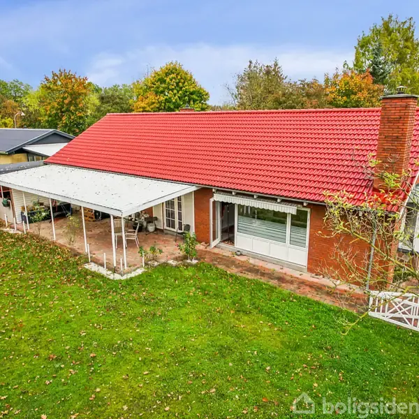 Rødt tegltagshus med terrasse og hvid udendørs overdækning omgivet af grøn have med nedfaldne blade. Området har flere huse og træer i efterårsfarver.