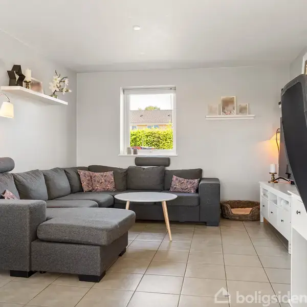 Grå hjørnesofa står i en stue med beige flisegulv. Sofaen har puder, og et rundt sofabord foran. Vindue og væghylder ses i baggrunden. Ingen tekst til stede.