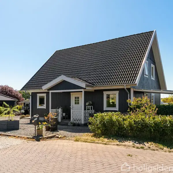 Et sort-hvidt hus med saddeltag står på en stille villavej, omgivet af velplejede buske og have. En brolagt indkørsel fører op til husets hovedindgang.