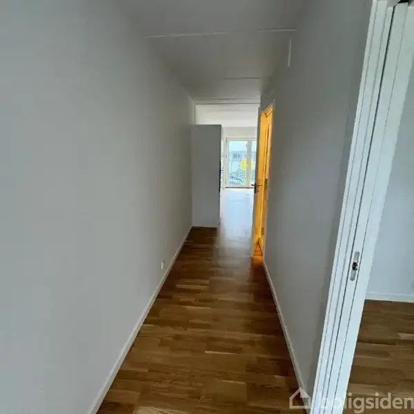 En lang gang med trægulv fører mod et oplyst rum for enden. På højre side er der en døråbning. Gangens vægge er hvide, og der er lys fra vinduer i baggrunden.