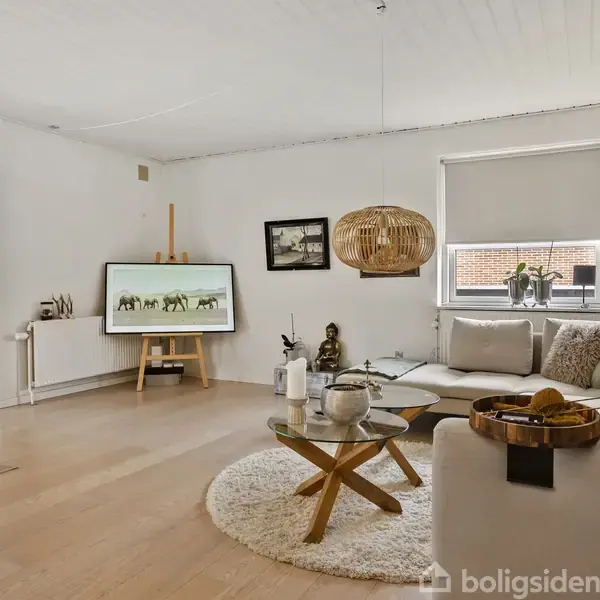 Stue med sofa og sofabord på rundt tæppe. Et TV står på et stativ, en lampe hænger ned fra loftet. Pejs i baggrunden, lysindfald fra vindue. Ingen tekst.